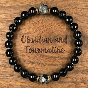 Puede incluir: Una pulsera de cuentas de obsidiana negra y turmalina. La pulsera presenta cuentas negras redondas y pulidas con una piedra central moteada y detalles dorados. Las palabras "Obsidian and Tourmaline" est&aacute;n grabadas en la superficie de madera.