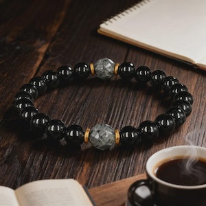 Puede incluir: Pulsera de cuentas negras con dos acentos de piedra gris facetada y espaciadores dorados. La pulsera se muestra sobre una superficie de madera oscura, con una taza de caf&eacute; y un cuaderno en el fondo. Una pieza de joyer&iacute;a.