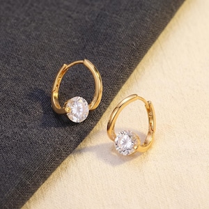 May include: A pair of gold hoop earrings featuring a polished metal finish and a single round, clear, faceted gemstone set at the base of each hoop. The earrings have a hinged closure mechanism and a curved, open design that holds the stone securely.
