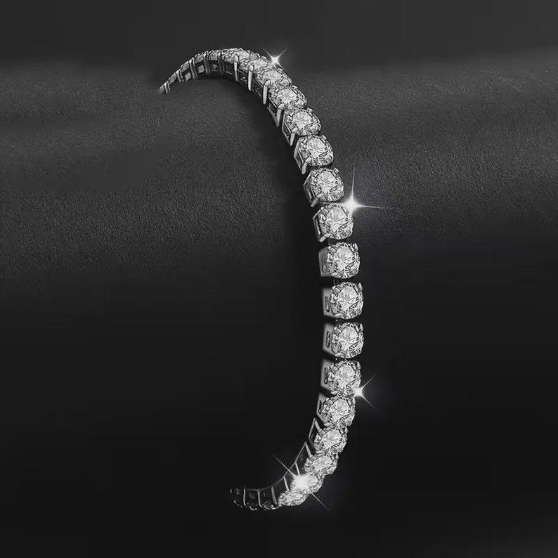 May include: A silver tennis bracelet with multiple round-cut, sparkling gemstones. The bracelet is curved, with the gemstones set closely together. The background is a dark, textured surface, creating a contrast with the bright bracelet.