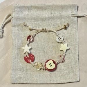May include: A handmade bracelet with button charms in various shapes, including stars, hearts, and flowers. The bracelet features a red apple button with a cream-colored center. The bracelet is on a beige drawstring bag.