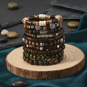 May include: A stack of wooden beaded bracelets in various shades of brown, green, and cream. The bracelets feature different bead sizes and textures, with some accented by metal and decorative elements. The bracelets are displayed on a round wooden slice.