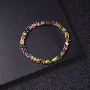 May include: A gold-toned bracelet with a row of square-cut, multi-colored gemstones. The gemstones are arranged in a rainbow spectrum, with a clasp closure. The bracelet is displayed on a dark surface.