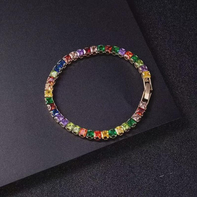 May include: A gold-toned bracelet with a row of square-cut, multi-colored gemstones. The gemstones are arranged in a rainbow spectrum, with a clasp closure. The bracelet is displayed on a dark surface.