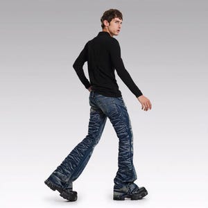 Op de afbeelding: Een persoon in een zwart shirt met lange mouwen en een blauwe spijkerbroek met een uniek, getextureerd ontwerp. De jeans heeft een uitlopende pijp en wordt gecombineerd met zwarte plateauzolen. De outfit staat tegen een neutrale achtergrond.