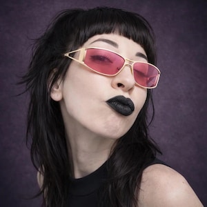 May include: Gold-framed sunglasses with pink lenses. The sunglasses have a rectangular shape and are worn by a person with black hair and black lipstick. The background is a solid purple color.