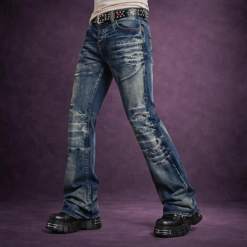 Op de afbeelding: Blauwe spijkerbroek met distressed details en een lichte flare aan de zoom. De jeans heeft vervaagde patches en een riem met studs. De persoon draagt zwarte plateau laarzen.