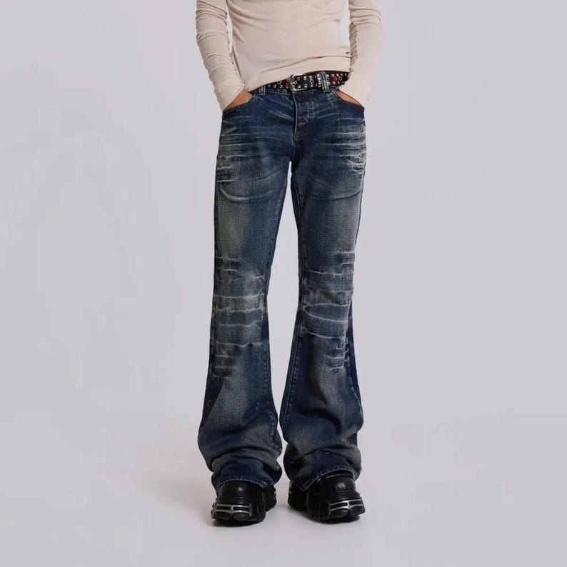 Op de afbeelding: Donkerblauwe denim jeans met een uitlopend pijp ontwerp. De jeans heeft distressed details en wordt gecombineerd met zwarte schoenen. Een riem met decoratieve studs is zichtbaar in de taille. De persoon draagt een lichtgekleurd shirt met lange mouwen.