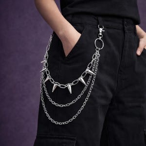 May include: Silver-toned metal chain with three tiers and cone-shaped spikes. The chain is attached to a pair of black pants with a silver clasp and ring. The background is a solid purple.