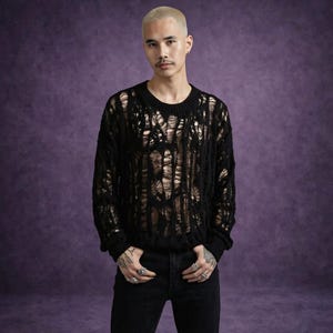 May include: Black, distressed knit sweater with a crew neck. The sweater has a shredded design, revealing the skin underneath. The model is wearing black jeans. The background is a solid purple color.
