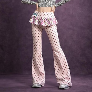 May include: Pink flared pants with a repeating pattern of dark red stars. The pants have a high waist with a ruffled trim featuring a plaid pattern. The pants are worn with silver shoes.