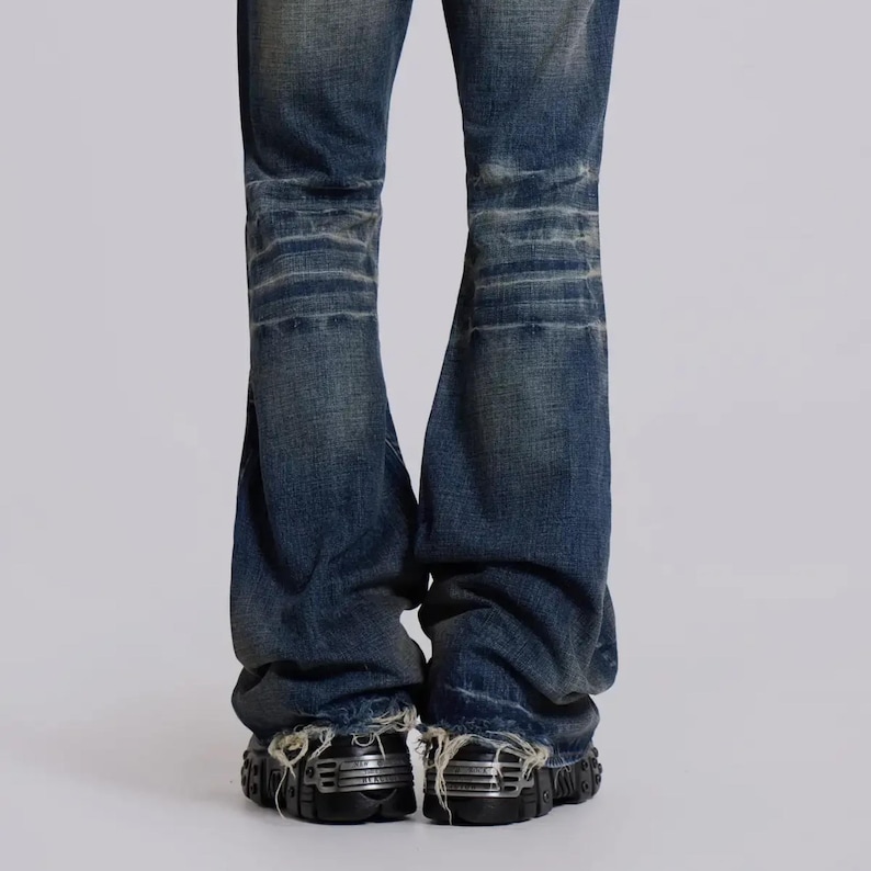 Op de afbeelding: Verbleekte blauwe denim jeans met distressed details. De jeans hebben een wijde beenopening en een versleten uiterlijk. De onderkant van de jeans is gerafeld. De jeans worden gecombineerd met zwarte schoenen met zilveren accenten.