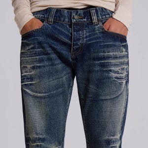 Op de afbeelding: Blauwe jeans van denim met een vervaagde en versleten uitstraling. De jeans hebben een knoopsluiting, een klassiek ontwerp met vijf zakken en een versleten look. De persoon draagt een shirt met lange mouwen.