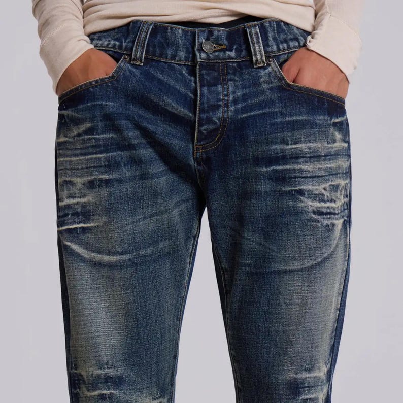 Op de afbeelding: Blauwe jeans van denim met een vervaagde en versleten uitstraling. De jeans hebben een knoopsluiting, een klassiek ontwerp met vijf zakken en een versleten look. De persoon draagt een shirt met lange mouwen.