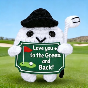 Op de afbeelding: Een wit gehaakt golfbal-personage met een zwarte hoed, die een witte golfclub en een groen bord vasthoudt. Op het bord staat "Love you to the Green and Back!" met een golfbal- en vlagillustratie. De achtergrond toont een golfbaan.