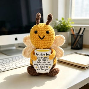 May include: A crocheted bee plush toy with yellow and brown yarn, black plastic eyes, and white wings. It holds a white sign shaped like a honey pot that reads: Positive Bee, Don't worry. Bee happy. I believe in you! Keep buzzing forward, no matter the challenge!