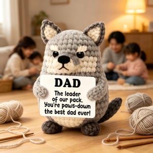 May include: A crocheted wolf figure in grey, white, and black, holding a sign that says "DAD, The leader of our pack. You're paws-down the best dad ever!" Yarn and knitting needles are visible. The figure is likely handmade.