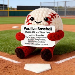 Könnte beinhalten: Ein gehäkelter Baseball-Charakter mit roten Nähten und Gliedmaßen, der ein Schild mit der Aufschrift "Positive Baseball" hält. Das Schild enthält inspirierenden Text über Anstrengung, Stärke und Selbstwertgefühl. Die Figur befindet sich auf einem Baseballfeld.