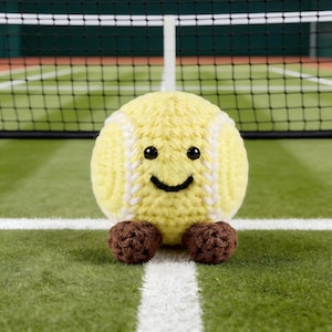 Peut inclure: Une balle de tennis jaune crochetée avec un visage souriant et des pieds marron. La balle de tennis a des détails de couture blancs et est positionnée sur un court de tennis vert avec un filet en arrière-plan.