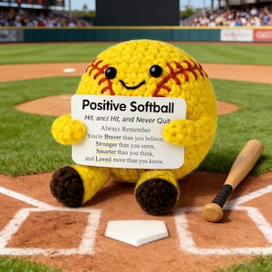 Könnte beinhalten: Ein gehäkelter gelber Softball-Charakter mit roten Nähten und einem lächelnden Gesicht, der ein Schild mit der Aufschrift "Positive Softball" hält. Ein Baseballschläger aus Holz liegt daneben auf einem Baseballfeld. Das Schild enthält inspirierenden Text.