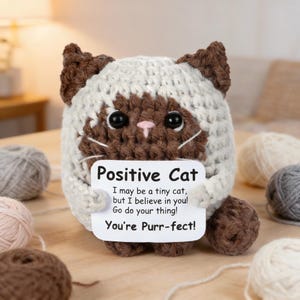 以下が含まれることがあります： 白と茶色の手編みの猫の置物。 「Positive Cat」と書かれたサインを持っています。 サインには「I may be a tiny cat, but I believe in you! Go do your thing! You're Purr-fect!」と書かれています。 背景には毛糸玉があります。