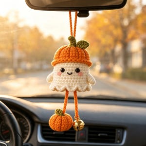 May include: A crocheted ghost ornament hanging from a rearview mirror. The ghost is white with black eyes, a small smile, and pink cheeks, wearing an orange pumpkin hat. Two orange yarn strands hang below, one with a small pumpkin and one with a bell.