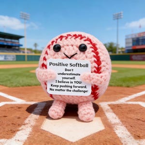 Könnte beinhalten: Ein gehäkelter Softball-Charakter mit einem lächelnden Gesicht, der ein Schild mit der Aufschrift "Positive Softball" hält. Das Schild ermutigt zum Selbstvertrauen und zur Ausdauer. Der Softball ist rosa mit roten Nähten, vor einem Baseballfeld.