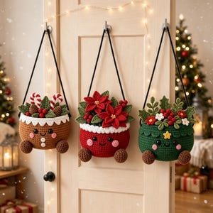 Op de afbeelding: Drie hangende gehaakte mandjes met lachende gezichtjes, elk met een eigen kerstthema. Het linker mandje is bruin met zuurstokjes, het middelste is rood met kerststerren en het rechter is groen met hulst en een klein geel sterretje.