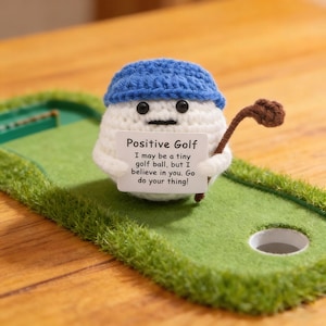 May include: A crocheted golf ball character wearing a blue hat and holding a golf club, positioned on a miniature green golf course. The character holds a sign that reads "Positive Golf" with an inspirational message. The golf course has a hole and artificial grass.