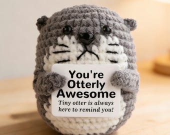 May include: A handmade crocheted otter in shades of grey and white, holding a sign with the words "You're Otterly Awesome." The sign also says "Tiny otter is always here to remind you!" The otter has black eyes and a black nose.