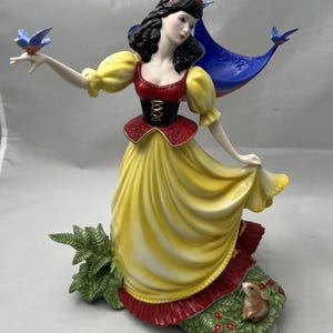 Peut inclure: Figurine en porcelaine d'une femme en robe jaune avec des accents rouges, tenant un oiseau bleu. La figurine a des cheveux noirs et un nœud rouge. Une structure bleue en forme de cape est derrière elle, et un petit lapin à ses pieds.