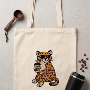 May include: A cream-colored canvas tote bag featuring a central embroidered patch of a leopard wearing black sunglasses and holding a coffee cup. The leopard has yellow fur with brown spots and is depicted in a seated position with a neutral expression.