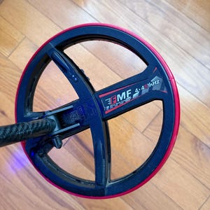 May include: A black and red metal detector coil with the text "EMF 4.45 kHz". The coil has a circular design with a red outer rim and a black interior. The detector is designed for finding metal objects.