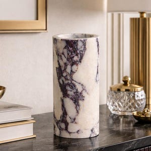 May include: A cylindrical vase crafted from white marble featuring prominent deep purple veining throughout its surface. The vase has a smooth, polished finish and a hollow center, standing upright as a decorative object with a distinct natural stone pattern.
