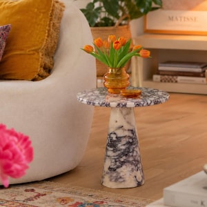 Peut inclure: Une table d'appoint ronde au motif de marbre blanc et violet foncé, dotée d'une base conique et d'un plateau plat. Un vase en verre ambré rempli de tulipes orange est posé sur la table, située à côté d'un fauteuil de couleur crème.