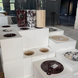 Puede incluir: Una colecci&oacute;n de recipientes cil&iacute;ndricos y bandejas circulares de piedra expuestos sobre bloques de espuma blancos. Los art&iacute;culos presentan varios patrones de piedra natural, incluyendo texturas de m&aacute;rmol rojo oscuro, blanco y negro, y travertino beige.