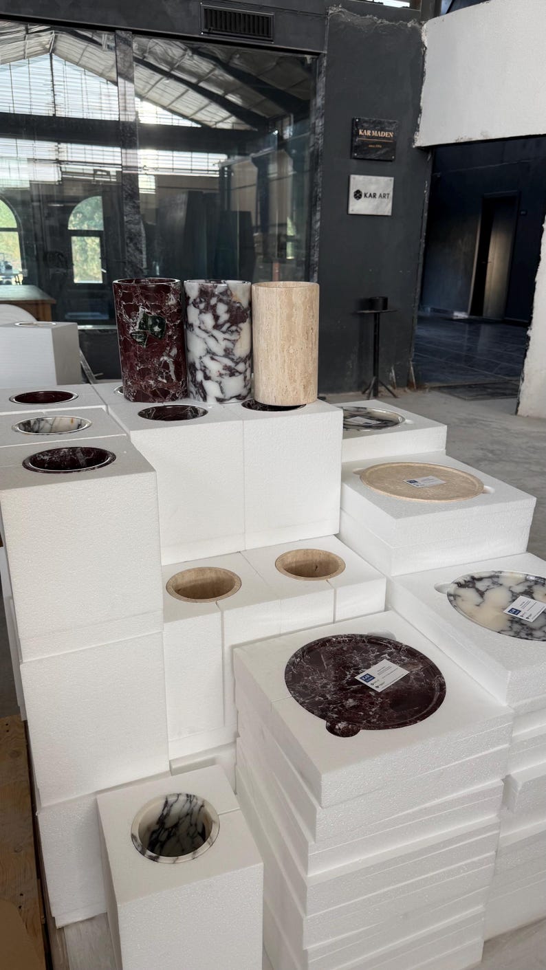 Puede incluir: Una colecci&oacute;n de recipientes cil&iacute;ndricos y bandejas circulares de piedra expuestos sobre bloques de espuma blancos. Los art&iacute;culos presentan varios patrones de piedra natural, incluyendo texturas de m&aacute;rmol rojo oscuro, blanco y negro, y travertino beige.