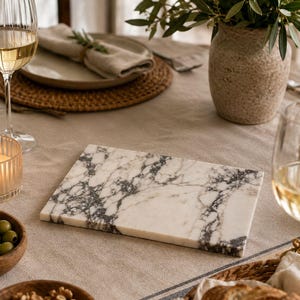 May include: A rectangular serving board made of white marble with prominent grey and black veining. The board has a smooth, polished surface and straight edges, positioned centrally on a neutral-colored fabric tablecloth among various dining items.