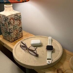 Puede incluir: Una bandeja redonda de hormig&oacute;n beige texturizado que contiene unas gafas de montura fina, un estuche de carga blanco para auriculares inal&aacute;mbricos y un reloj inteligente con correa de silicona blanca. La bandeja tiene una superficie porosa.