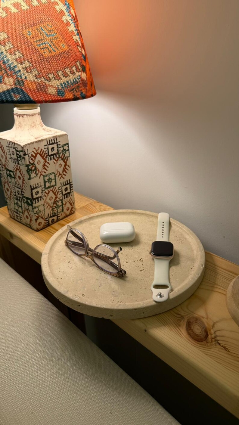Puede incluir: Una bandeja redonda de hormig&oacute;n beige texturizado que contiene unas gafas de montura fina, un estuche de carga blanco para auriculares inal&aacute;mbricos y un reloj inteligente con correa de silicona blanca. La bandeja tiene una superficie porosa.