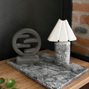Puede incluir: Un quemador de incienso circular gris oscuro con niveles horizontales y una pequeña lámpara de mesa con pantalla blanca plisada sobre una base cilíndrica gris moteada, ambos sobre una losa rectangular de mármol veteado gris y blanco.
