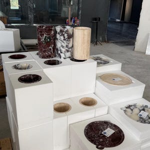 Puede incluir: Una colecci&oacute;n de recipientes cil&iacute;ndricos y bandejas circulares de piedra expuestos sobre bloques de espuma blancos. Los art&iacute;culos presentan varios patrones de piedra natural en tonos rojo oscuro, blanco y negro, y beige. Algunas piezas tienen etiquetas.