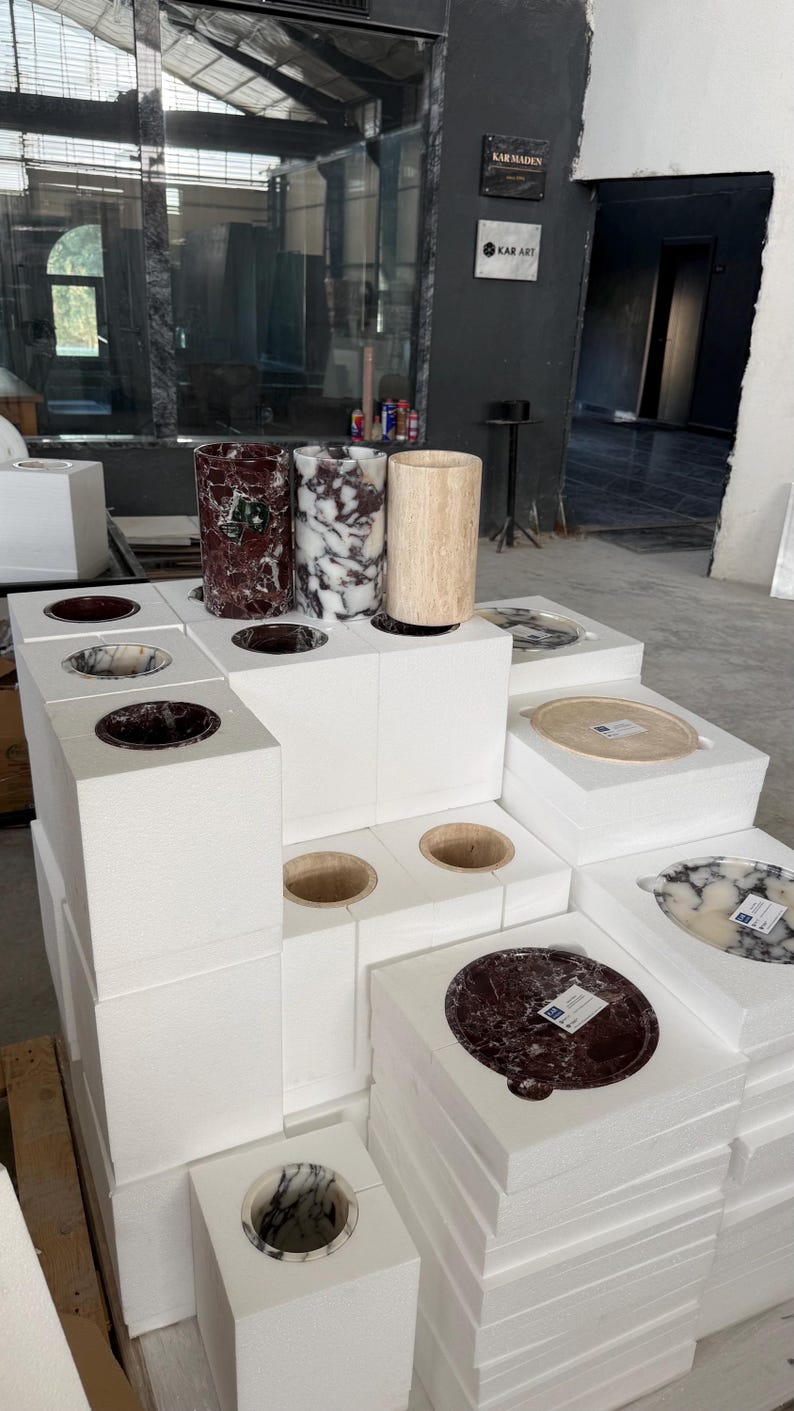 Puede incluir: Una colecci&oacute;n de recipientes cil&iacute;ndricos y bandejas circulares de piedra expuestos sobre bloques de espuma blancos. Los art&iacute;culos presentan varios patrones de piedra natural en tonos rojo oscuro, blanco y negro, y beige. Algunas piezas tienen etiquetas.