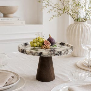 May include: A circular cake stand featuring a white and black marbled stone top supported by a dark brown, conical wooden base. The stand holds a small cluster of green grapes, a halved purple fig, and a few shelled walnuts on its surface.