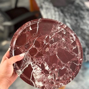 May include: A circular tray crafted from deep burgundy red marble featuring prominent white and light pink veining throughout the surface. The tray has a smooth, polished finish and a raised edge that runs along the entire circumference of the piece.