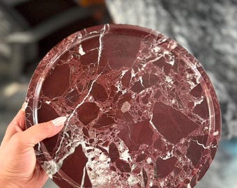 Könnte beinhalten: Ein kreisförmiges Tablett aus tief burgunderrotem Marmor mit markanter weißer und hellrosa Maserung auf der gesamten Oberfläche. Das Tablett hat eine glatte, polierte Oberfläche und einen erhöhten Rand, der entlang des gesamten Umfangs verläuft.