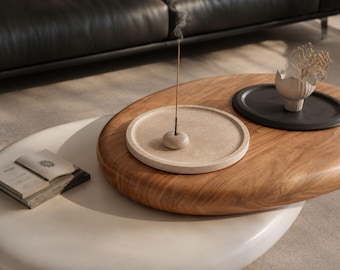 Puede incluir: Una mesa de centro ovalada de madera marrón claro con una bandeja circular beige moteada que contiene un pequeño porta-incienso y una varilla de incienso encendida, junto a una bandeja circular negra con un jarrón blanco texturizado.