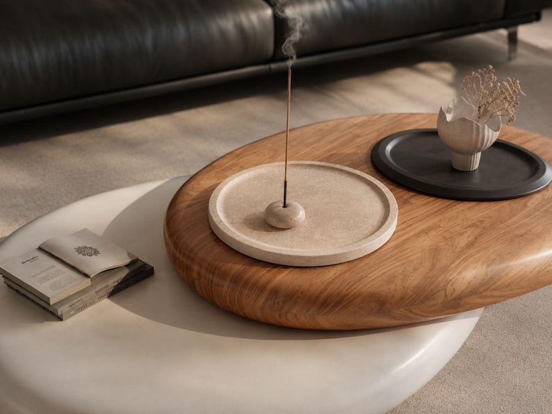 Puede incluir: Una mesa de centro ovalada de madera marr&oacute;n claro con una bandeja circular beige moteada que contiene un peque&ntilde;o porta-incienso y una varilla de incienso encendida, junto a una bandeja circular negra con un jarr&oacute;n blanco texturizado.