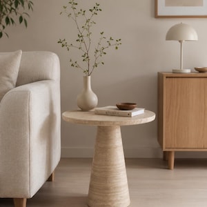 May include: A round side table crafted from beige travertine stone with a textured, porous surface and a wide, conical pedestal base. The table surface holds a smooth, light-colored ceramic vase with green branches, a small wooden bowl, and a book.
