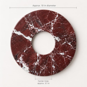 May include: A circular red marble trivet with white veining, featuring a center hole. The text indicates an approximate 10-inch diameter and a center hole of approximately 3.1 inches. The surface displays natural stone patterns and textures.